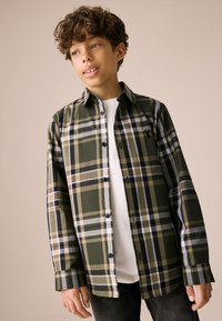 Chemise à carreaux avec des couleurs vert, beige et noir, design boutonné et manches longues, portée sur une chemise blanche. Texture de tissu lisse.