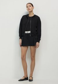 Haut en molleton noir coupe courte avec fermeture éclair à l'avant et poches latérales, associé à une jupe plissée noire. Des chaussures plates noires complètent la tenue.