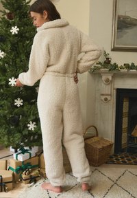 Cremefarbenes, flauschiges Overall mit Kragen und elastischem Bund, lange Ärmel und schmal zulaufende Beine, vor einem dekorierten Weihnachtsbaum.
