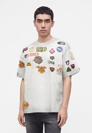 Jeune homme aux cheveux noirs et courts porte un t-shirt oversize gris clair décoré de multiples patchs brodés colorés et un jean foncé.