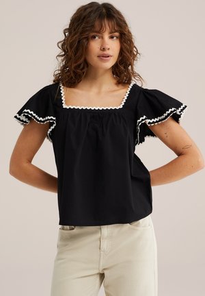 Žena nosi čiernu blúzku s bielym zvlněným lemom na štvorcovom výstrihu a s volánovými rukávmi, v kombinácii so svetlo béžovými nohavicami.