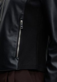 Gros plan sur une veste noire avec une poche verticale zippée argentée sur un panneau en tissu lisse et mat.