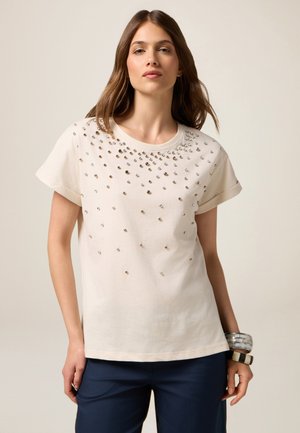 T-shirt di cotone beige chiaro con maniche corte, colletto rotondo, decorato con cristalli di vetro argentati di diverse dimensioni sparsi sulla parte anteriore.