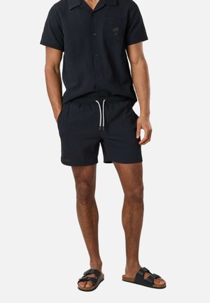 Homme portant une chemise noire à manches courtes boutonnée, un short noir à cordon de serrage et des sandales noires à double bride sur un fond uni.