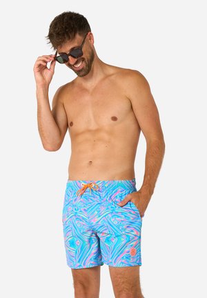 Hemdloser Mann lächelt und richtet seine Sonnenbrille, trägt bunte blaue und rosa gemusterte Badeshorts mit orangefarbenem Kordelzug, Hand in der Tasche.