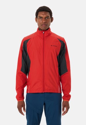 Uomo che indossa una giacca Vaude rossa e nera con zip e pantaloni blu, in piedi davanti a uno sfondo chiaro e uniforme, rivolto verso l'obiettivo.