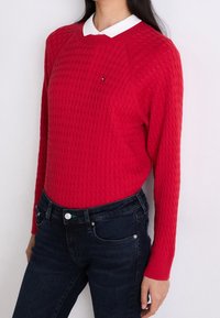 Suéter rojo de punto con textura acanalada, cuello redondo y cuello de camisa blanco, combinado con jeans azul oscuro. Presente un pequeño emblema de logo.