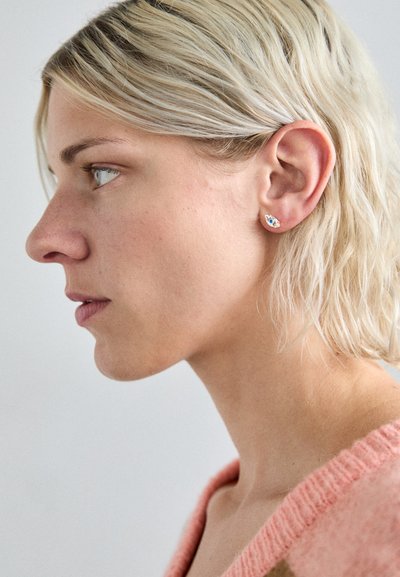 Pendientes de metal dorado con un diseño de ojo azul y blanco, con un acabado suave y en forma de lágrima, sujetos a una oreja.