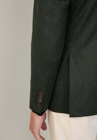 Mörkgrön ullblazer med subtil struktur, featuring armbågsfickor, fyra bruna knappar på ärmslutet och en skräddarsydd passform.