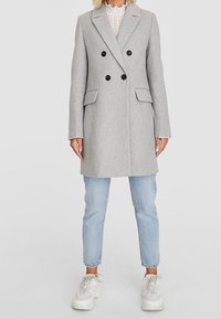 Cappotto grigio in lana doppiopetto con bottoni neri, tasche laterali e rever a punta. Indossato sopra jeans azzurri chiari e scarpe da ginnastica bianche spesse.