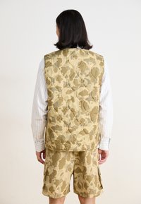 Tan camouflagemönstrad vadderad väst med rundade kanter och stickat mönster, matchande shorts. Randig tröja under, bakifrån.