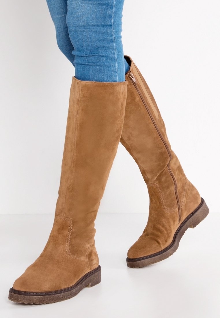 Person som bär knähöga, beige mockastövlar med sidodragkedja och blå skinny jeans mot vit bakgrund.