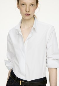 White button-up shirt with rolled sleeves, made of smooth fabric. Features a classic collar and is paired with dark denim and a black belt.