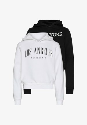 Fehér kapucnis pulóver, rajta fekete betűkkel "LOS ANGELES" és "CALIFORNIA" felirattal, világoskék farmernadrággal és bézs sportcipővel párosítva. Göndör haj.