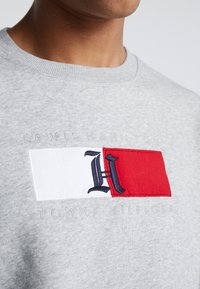 Gros plan d'un sweat-shirt gris avec un écusson rouge et blanc affichant un "H" marine et le texte en relief "Lewis Hamilton" et "Tommy Hilfiger".