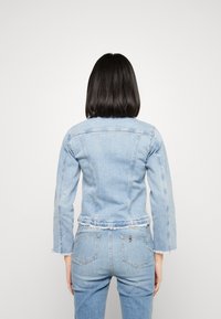 Mujer con cabello oscuro y lacio, vistiendo una chaqueta de mezclilla azul claro y jeans a juego, de pie de espaldas contra un fondo blanco liso.