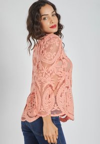Blusa de encaje melocotón con bordado floral, mangas largas transparentes y dobladillo con forma de concha. Combinada con vaqueros oscuros, exhibe un escote redondo.