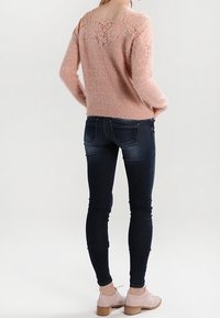 Rosa, kuscheliger Pullover mit Spitzenakzenten auf der Rückseite, kombiniert mit dunklen Skinny-Jeans und hellrosa Blockabsatzschuhen.