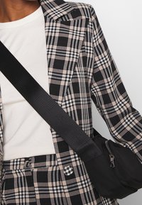 Černobílý kostkovaný blejzr přes bílé tričko, doplněný černou křížovou taškou se zipem a nastavitelným popruhem.