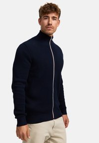 Suéter de punto azul marino con cremallera y cuello alto, que presenta una tela texturizada, puños y dobladillo acanalados, y un detalle de cremallera en contraste.