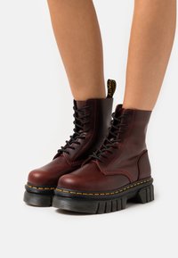 Botas de tobillo de cuero marrón con cordones negros, suela de goma gruesa, costura amarilla y una etiqueta en bucle en la parte posterior.