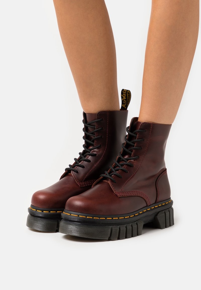 Botas de tobillo de cuero marrón con cordones negros, suela de goma gruesa, costura amarilla y una etiqueta en bucle en la parte posterior.