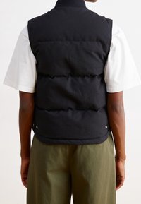 Černá zateplená vesta s vysokým límcem, s prošívaným designem a knoflíkovými uzávěry po stranách. Nositelná přes bílou košili a olivové kalhoty.