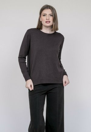 Femme aux cheveux raides mi-longs, portant un pull à manches longues foncé et un pantalon large noir, devant un fond uni.