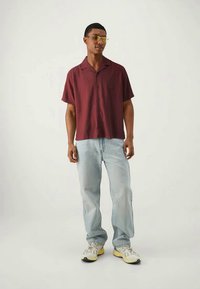 Camisa de boton corto de manga corta burdeos, corte relajado; jeans de mezclilla azul claro; zapatillas deportivas blancas y amarillas; gafas de sol amarillas transparentes.