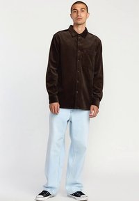 Bruine corduroy blouse met lange mouwen, knoopsluiting aan de voorkant, enkele borstzak. Gecombineerd met lichtblauwe jeans met rechte pijpen en zwarte sneakers.