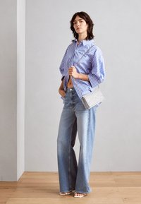 Chemise oversize à rayures bleues et blanches, jeans larges à lavage clair, sac à main argenté métallisé, et sandales à talons. Textures de tissu lisses visibles.