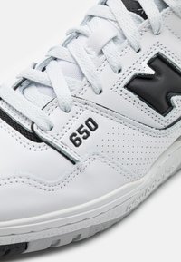 Vita lädersneakers med svarta detaljer, perforerade inslag, snörning och modellnummer 650 på sidan. Gummisula synlig.