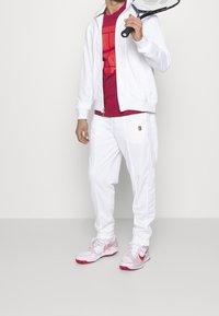 Witte sportjack met rits, rode T-shirt met patroon, witte broek met logo en wit-roze sportschoenen. Houdt een racket vast.