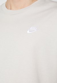 Ljusgrå bomullströja med rund halsringning och en liten vit broderad Nike-logotyp på bröstet. Slät textur.