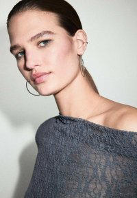 Top gri din dantelă cu design off-shoulder, având un model texturat. Modelul poartă cercei mari din argint, evidențiind un machiaj natural și părul drept.