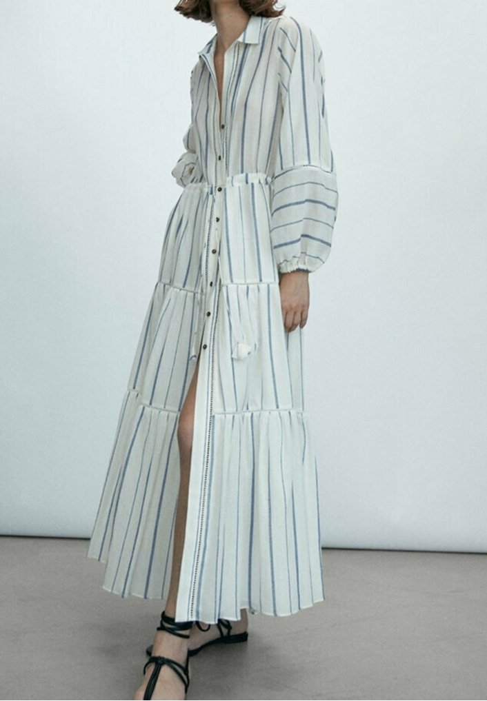 Femme portant une longue robe blanche à rayures bleues verticales, manches longues bouffantes, boutons sur le devant et sandales noires à lacets.