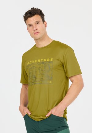 Mann trägt olivgrünes T-Shirt mit der Aufschrift "ADVENTURE" und topografischem Kartenmuster, Hände in den Taschen, steht vor einem einfarbigen weißen Hintergrund.