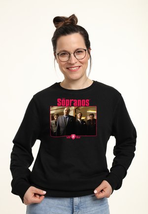 Kobieta w okularach i czarnej bluzie z wizerunkiem i logo obsady serialu The Sopranos, uśmiechająca się na jednolitym jasnym tle.