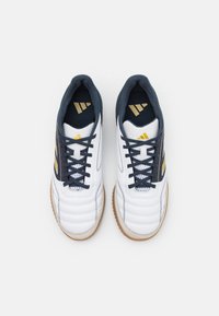 Par de zapatos de fútbol sala blancos con cordones negros, rayas doradas y suelas de goma vista desde arriba sobre un fondo blanco.