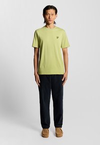 Lys grøn bomulds T-shirt med korte ærmer og logo på brystet, parret med mørk navy bukser og brune ruskinds slip-on sko. Enkel design.