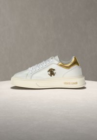 Λευκό χαμηλό sneaker με χρυσή λεπτομέρεια στη φτέρνα, χρυσό λογότυπο στο πλάι και κείμενο "roberto cavalli" στη σόλα πάνω σε ουδέτερο φόντο.
