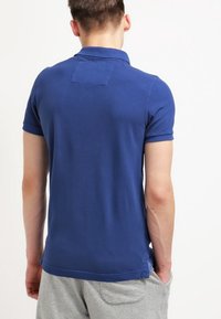 Mörkblå pikétröja med korta ärmar, platt krage och en patch på övre ryggen. Slät bomullstextur, avslappnad passform.