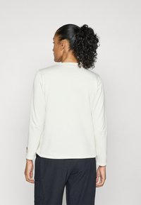 Camisa de manga larga en tejido texturizado de color crema claro con un cuello redondo, que presenta un pequeño logotipo en la parte inferior de la manga izquierda.