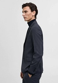 Dark gray suit with a subtle check pattern, featuring a notched lapel and two front pockets, paired with a black turtleneck underneath.