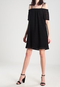 Schwarzes, schulterfreies Kleid mit kurzen Ärmeln, das ein strukturiertes Muster aufweist. Kombiniert mit schwarzen, hochhackigen Sandalen.