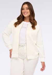 Bílá zipovací bunda s texturovaným, diamantovým vzorem na přední části a jednobarevnými rukávy; spárována s bílou košilí a odpovídajícími kalhotami.