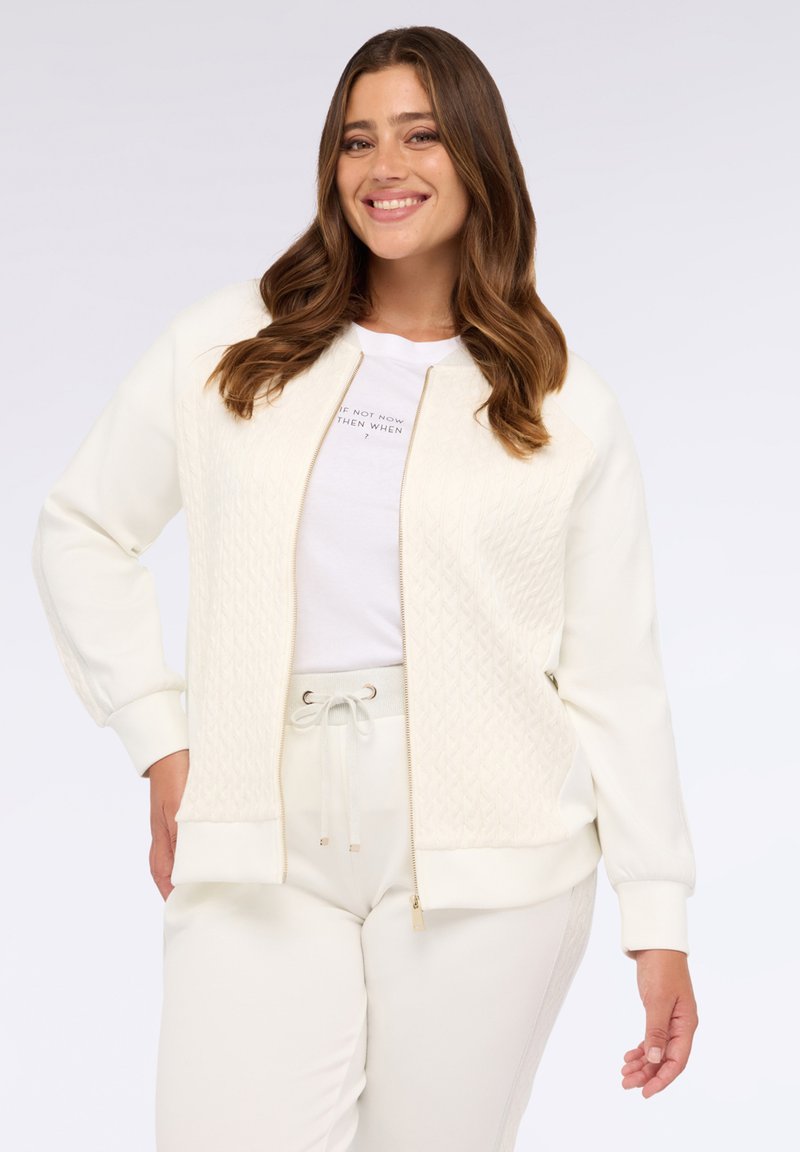 Bílá zipovací bunda s texturovaným, diamantovým vzorem na přední části a jednobarevnými rukávy; spárována s bílou košilí a odpovídajícími kalhotami.