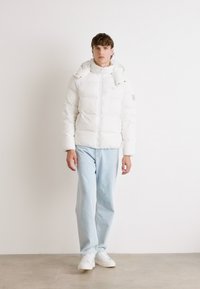 Vit vadderad jacka med dragkedja och huva; bärs över ljusblå jeans och vita sneakers; jämn textur med horisontell sömnad.
