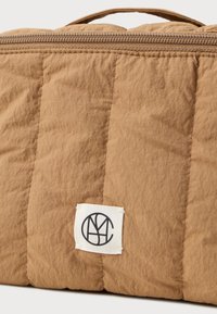 Φως καφέ τσάντα από επένδυση με μαύρη και λευκή κυκλική ετικέτα λογότυπου κοντά στο κάτω μέρος και ορατό φερμουάρ στην κορυφή.