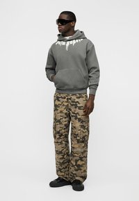 Homme portant des lunettes de soleil noires, un sweat à capuche gris avec texte blanc, un pantalon cargo camouflage et des chaussures noires, debout les mains dans les poches.